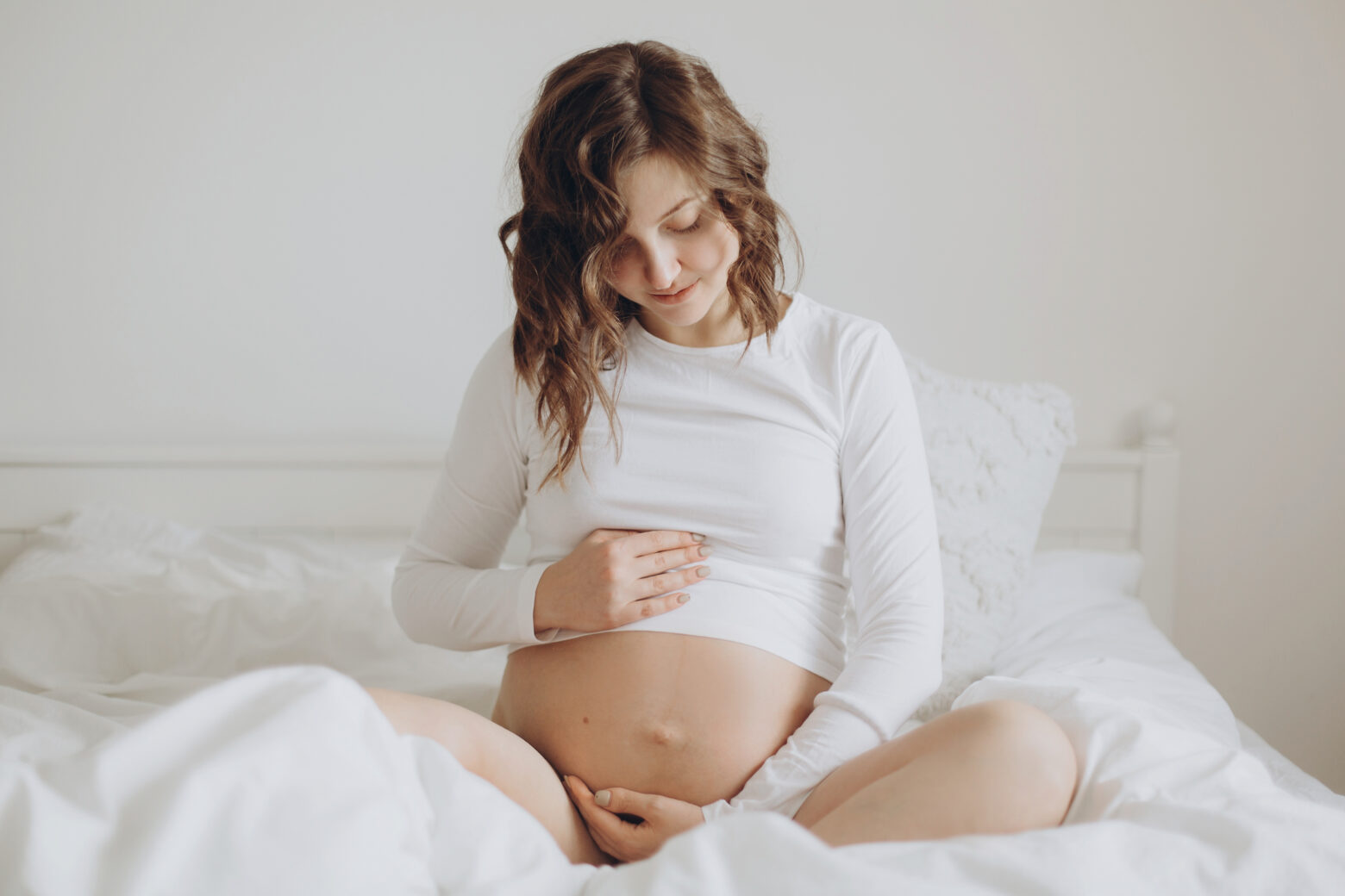 Happy pregnant woman in white holding belly bump and relaxing on white bed at home. Stylish pregnant mom hugging belly with love and care, waiting for baby. Motherhood concept ,maternity time